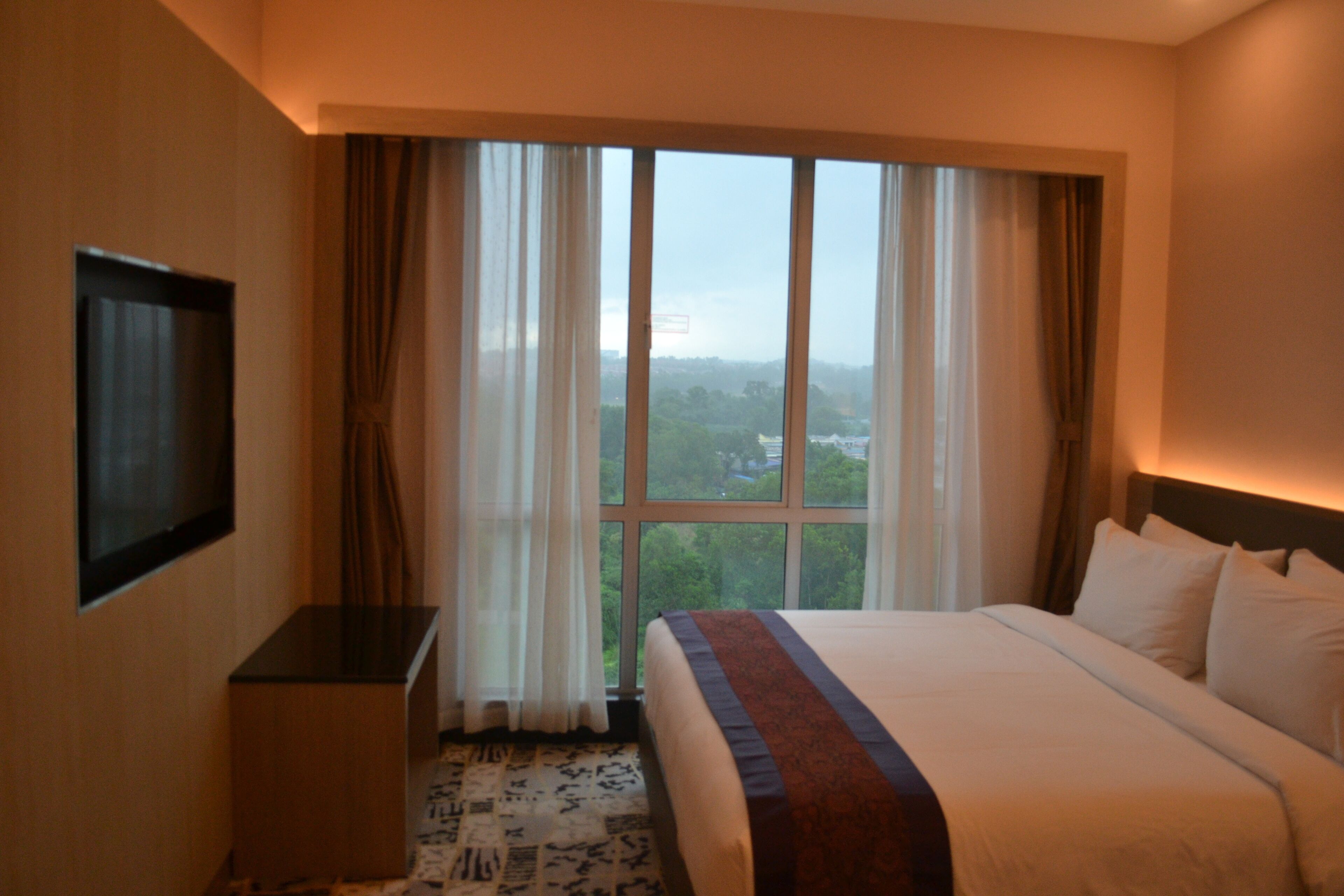 A hotel room with a large bed, a flat-screen TV mounted on the wall, and a window with curtains.