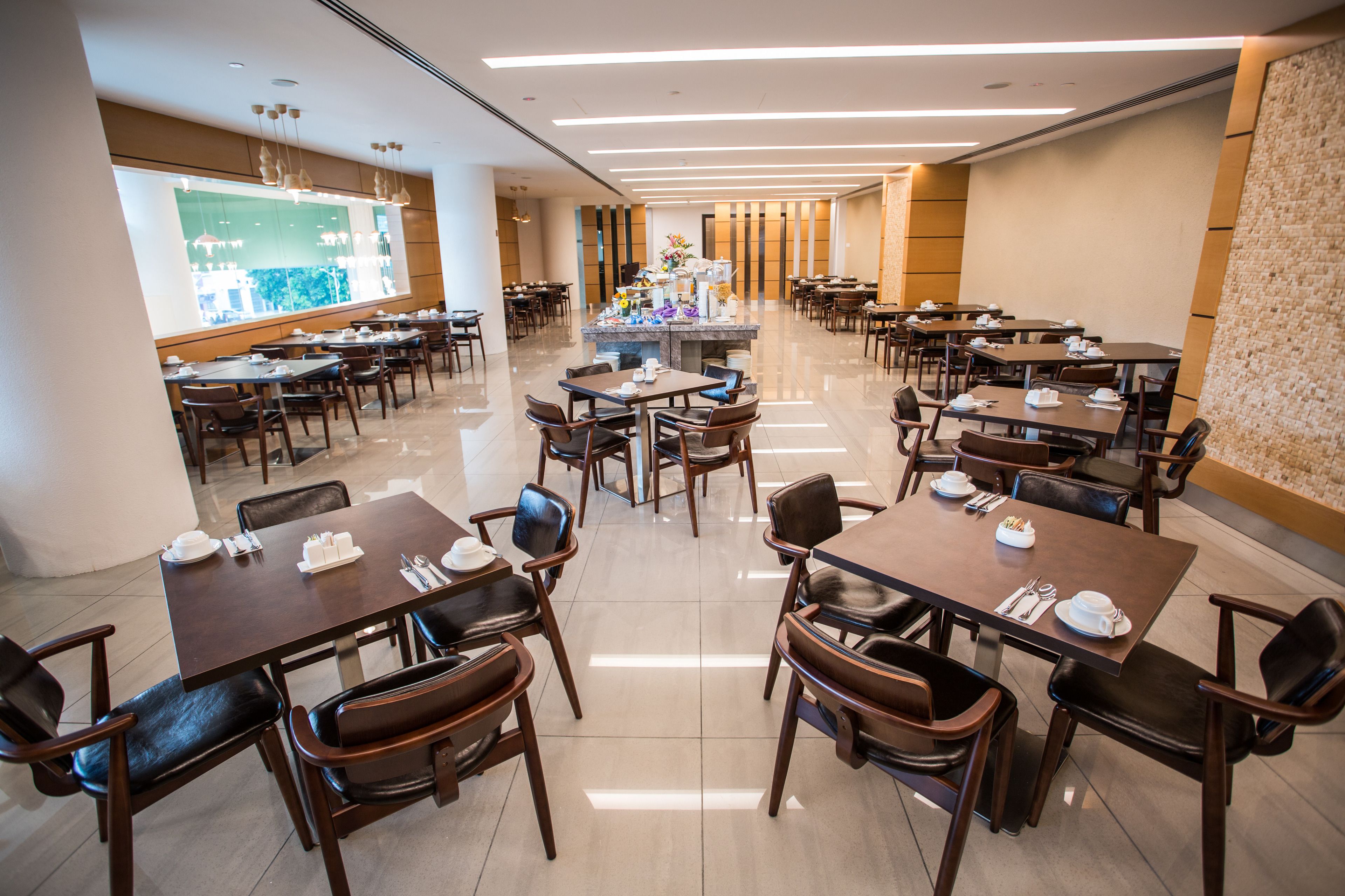 A spacious hotel dining area with multiple tables and chairs, a central decorative island, and a large window with a view.