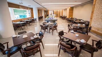 A spacious hotel dining area with multiple tables and chairs, a central decorative island, and a large window with a view.