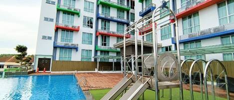 A swimming pool with a slide, surrounded by a building with multiple balconies and a green roof.