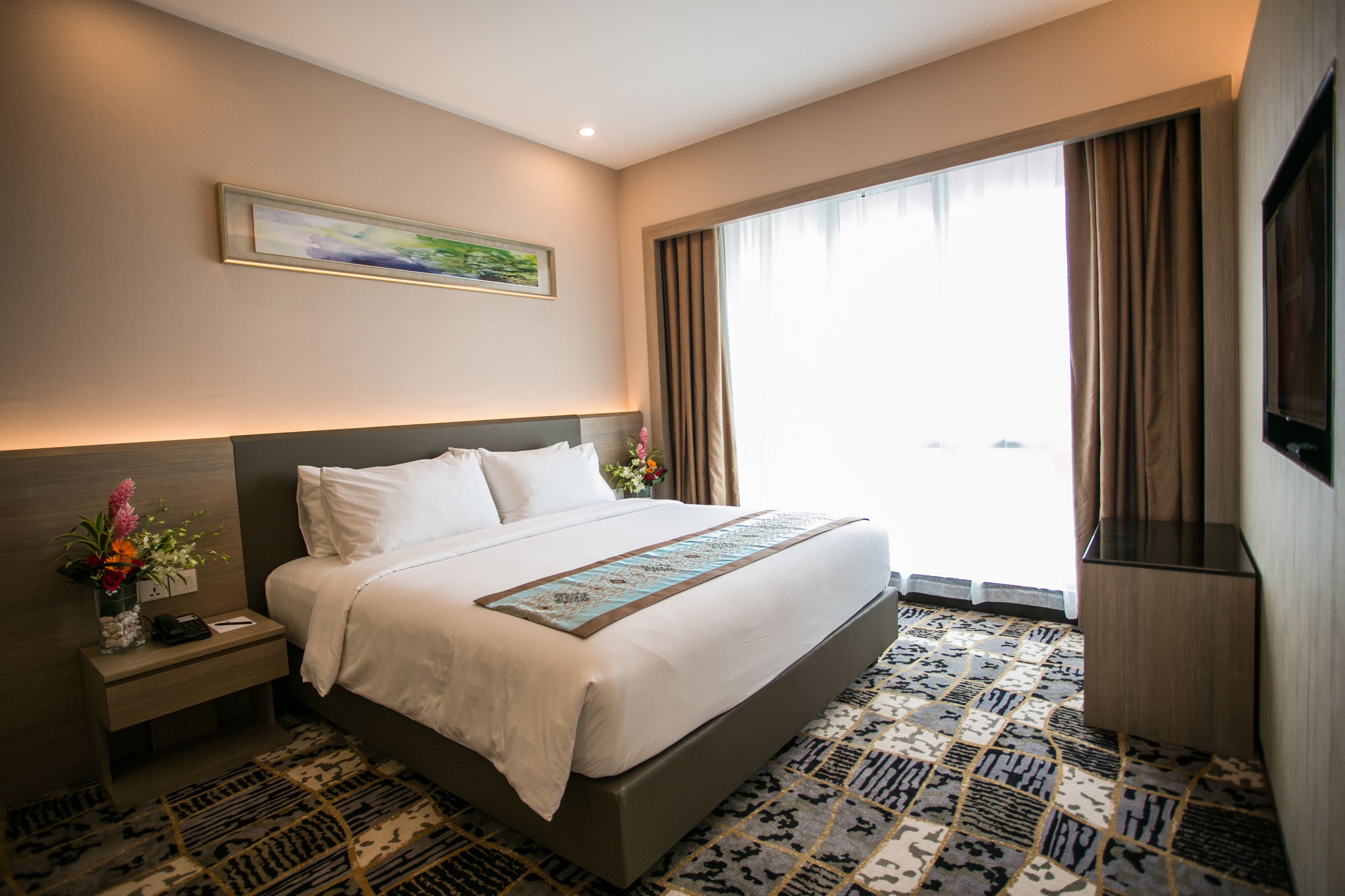 A hotel room with a large bed, a nightstand with a phone and a vase of flowers, a painting on the wall, and a large window with curtains.