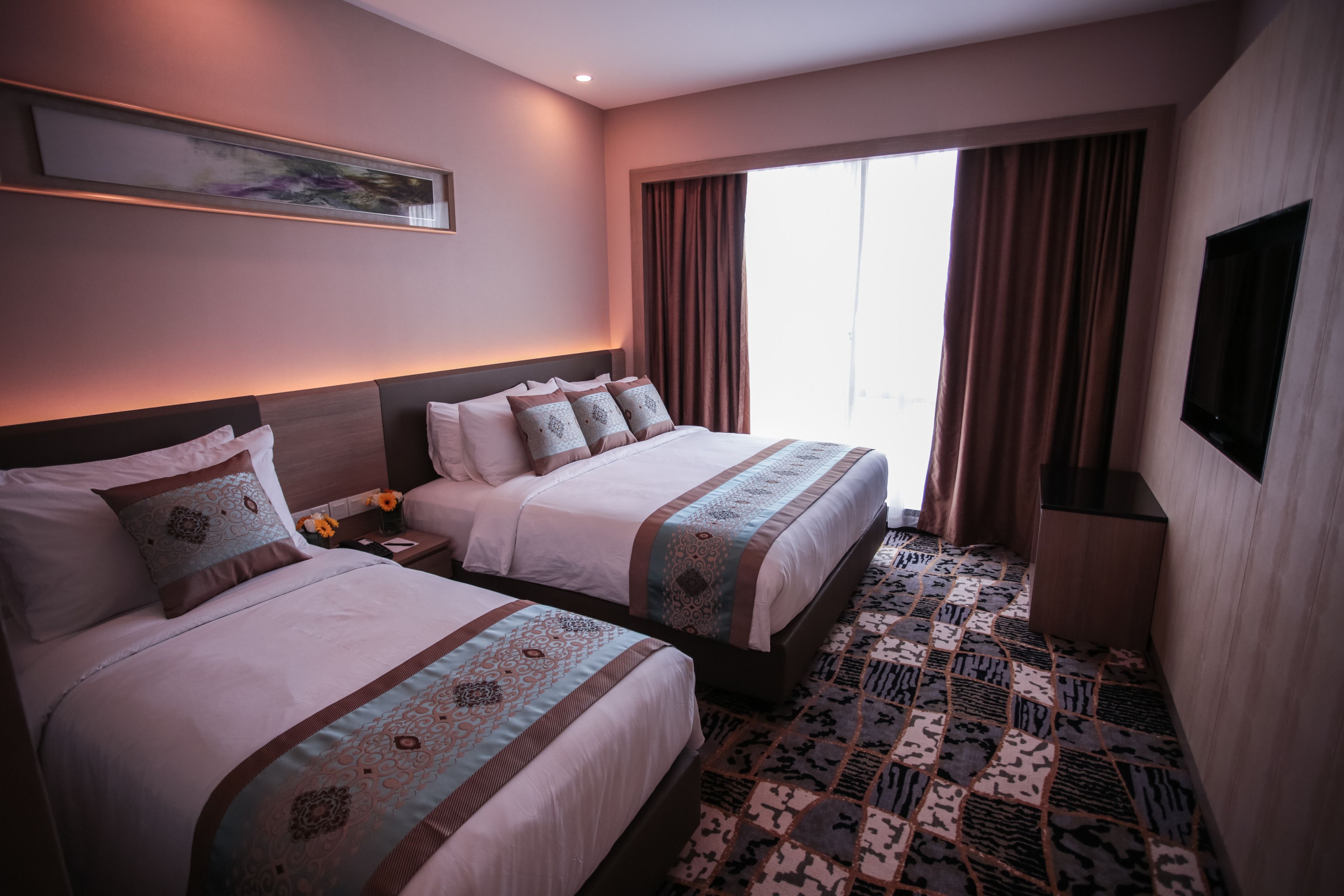 A hotel room with two beds, a large window with curtains, a flat-screen TV mounted on the wall, and a patterned carpet.