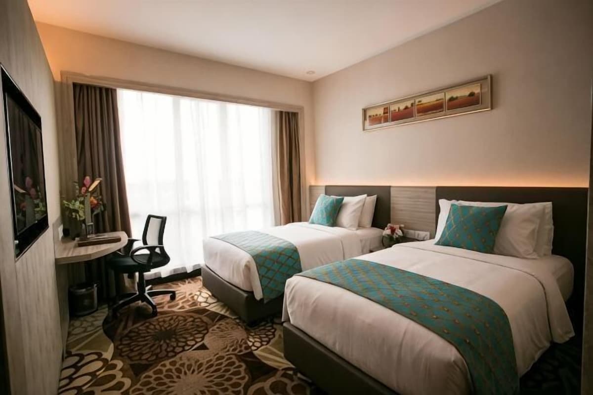 Habitación de hotel con dos camas, un escritorio con silla, una mesita pequeña y un ventanal con cortinas.