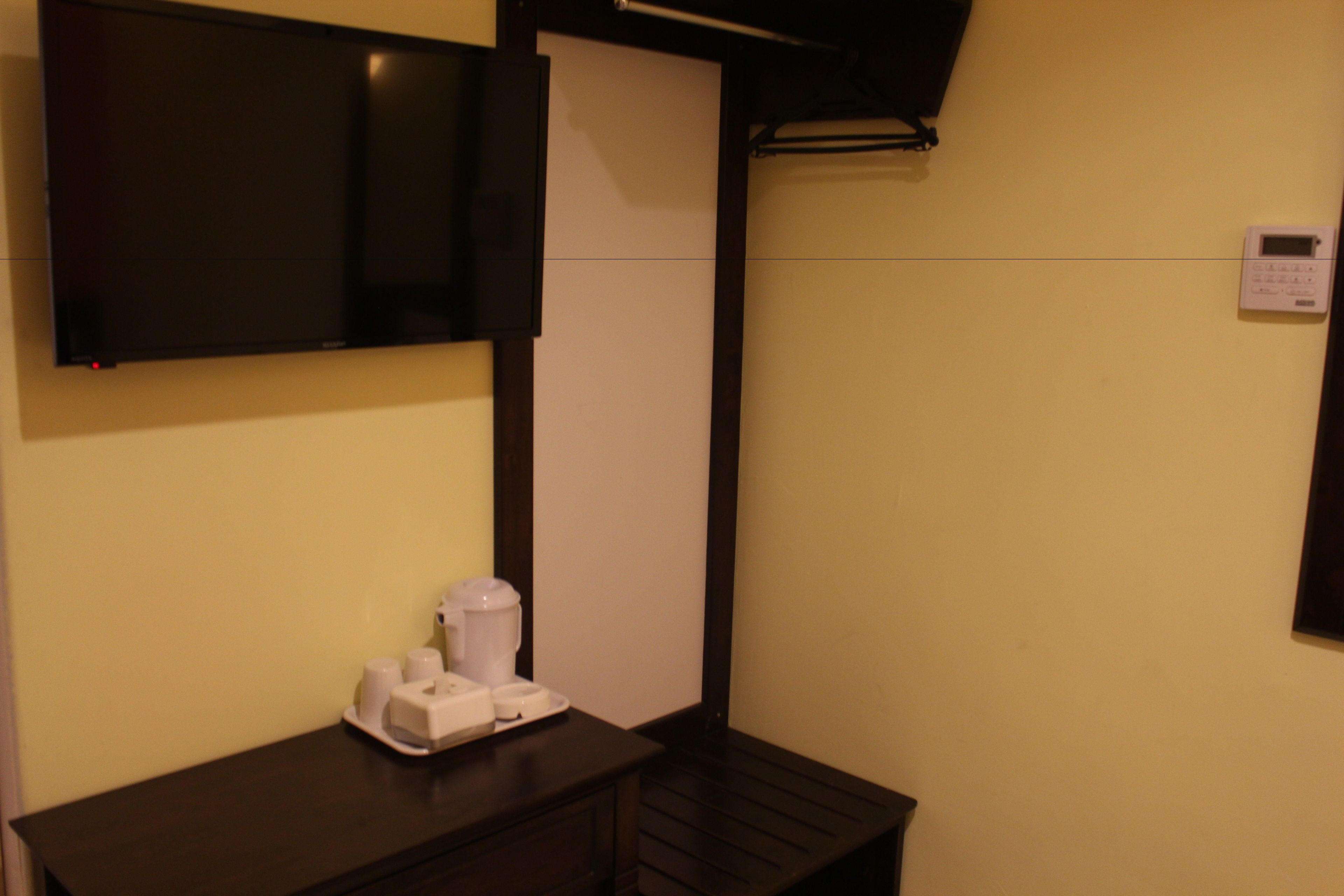A hotel room with a flat-screen TV mounted on the wall, a coffee maker on a wooden table, and a thermostat on the wall.