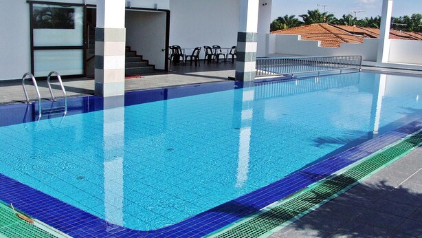 Una piscina con agua azul cristalina, un solárium con baldosas y una zona de estar techada con sillas.