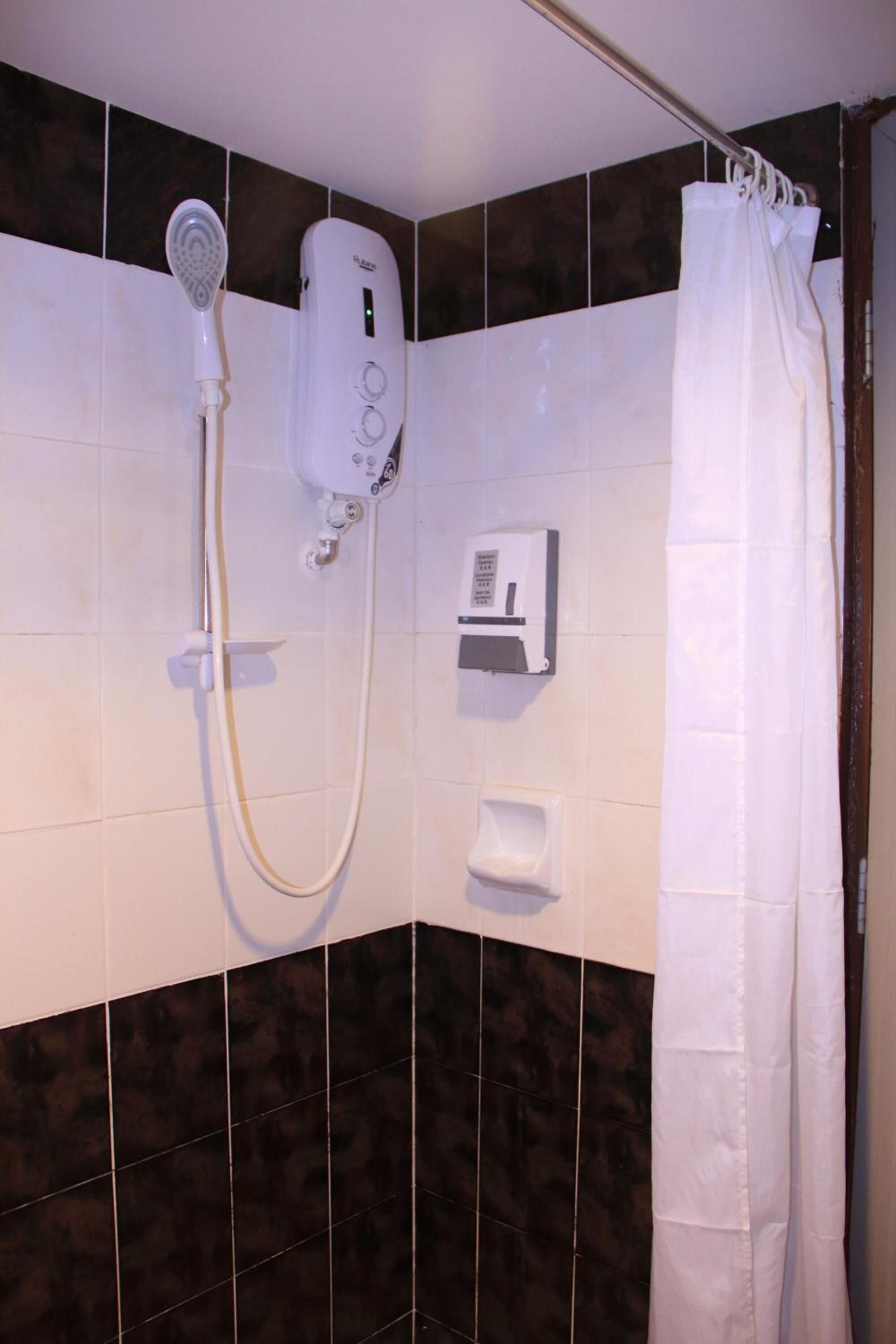 A shower area with a handheld showerhead, a wall-mounted control panel, a soap dispenser, and a white shower curtain.