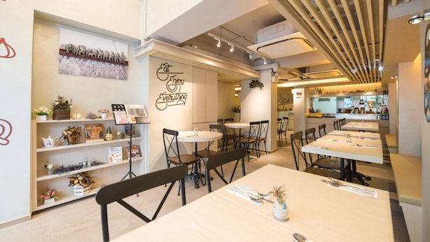 Interior moderno de restaurante com várias mesas e cadeiras, um quadro de menu fixado na parede e prateleiras decorativas com flores e livros.