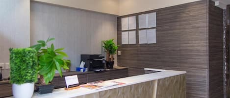 A reception desk with a modern design, featuring a textured wall, a flat-screen TV, and a printer. There are potted plants on the desk, and a sign with information.