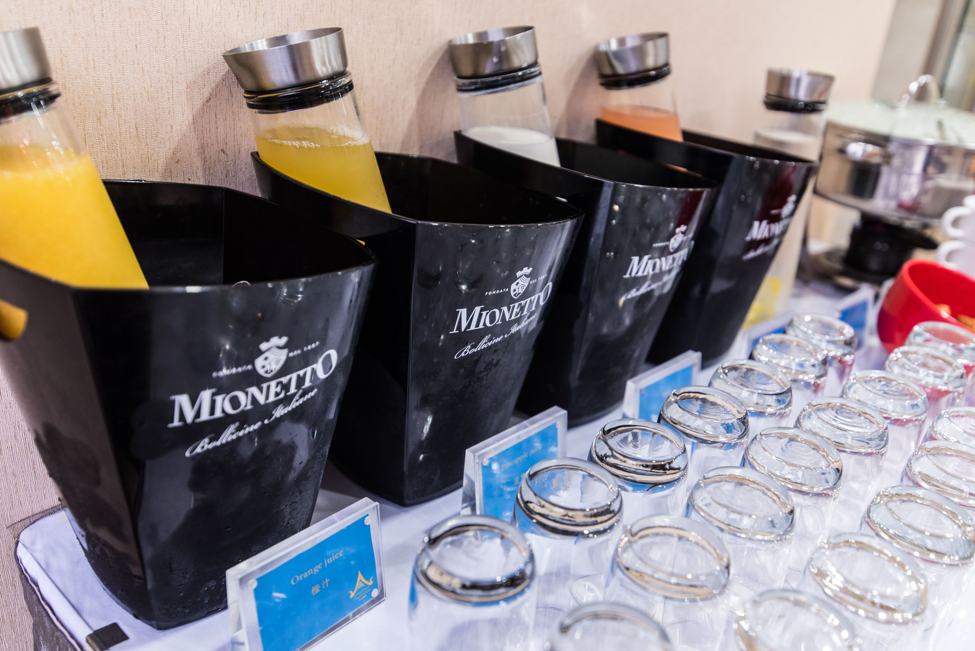 A selection of Mionetto branded drink dispensers with various colored liquids, and a display of clear glass tumblers on a table.