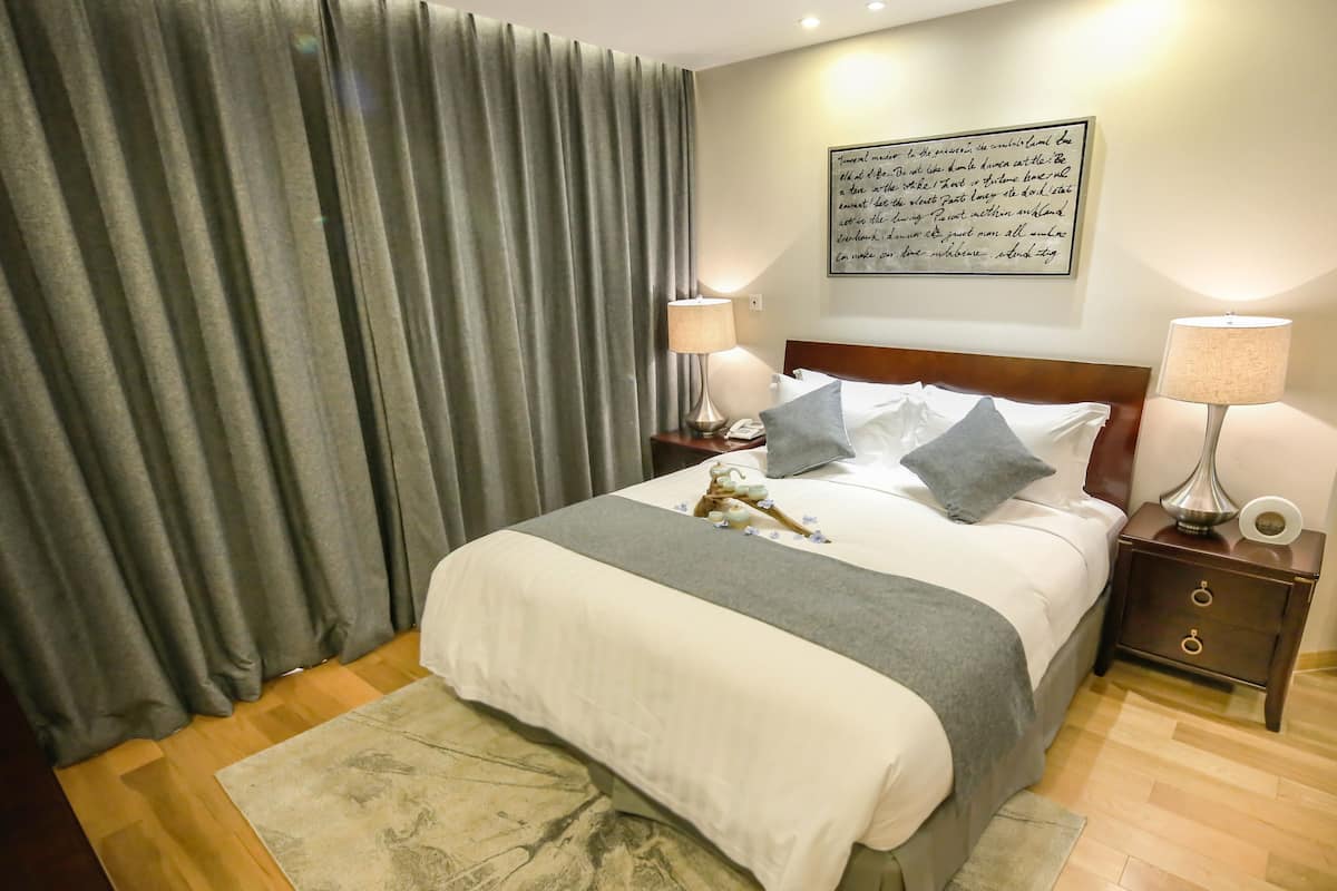 A neatly made bed with pillows and a blanket, a bedside table with a lamp, a wall with a framed quote, and a large window with curtains.
