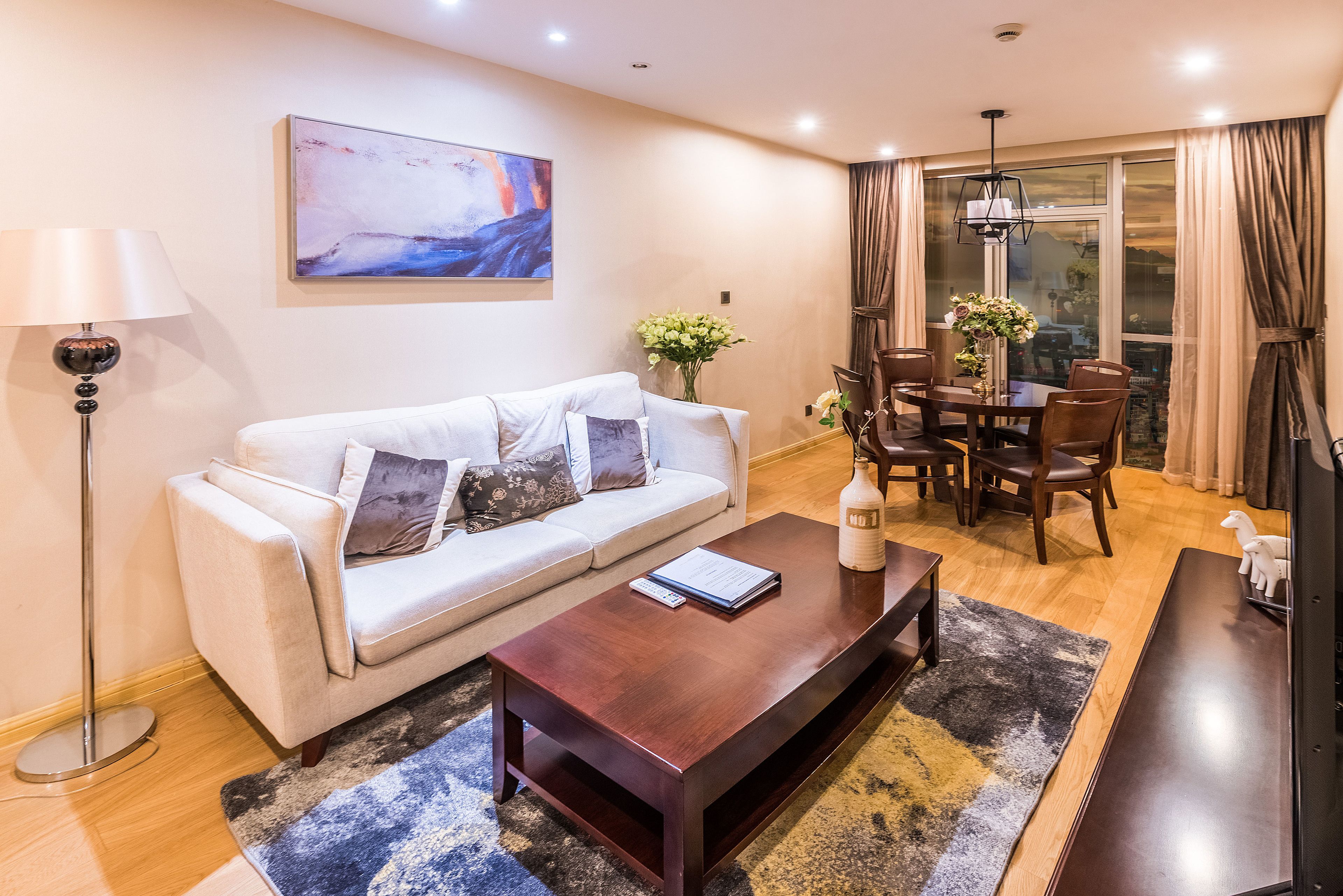 A modern living room with a white sofa, a coffee table, a dining table with chairs, and a large window with curtains.