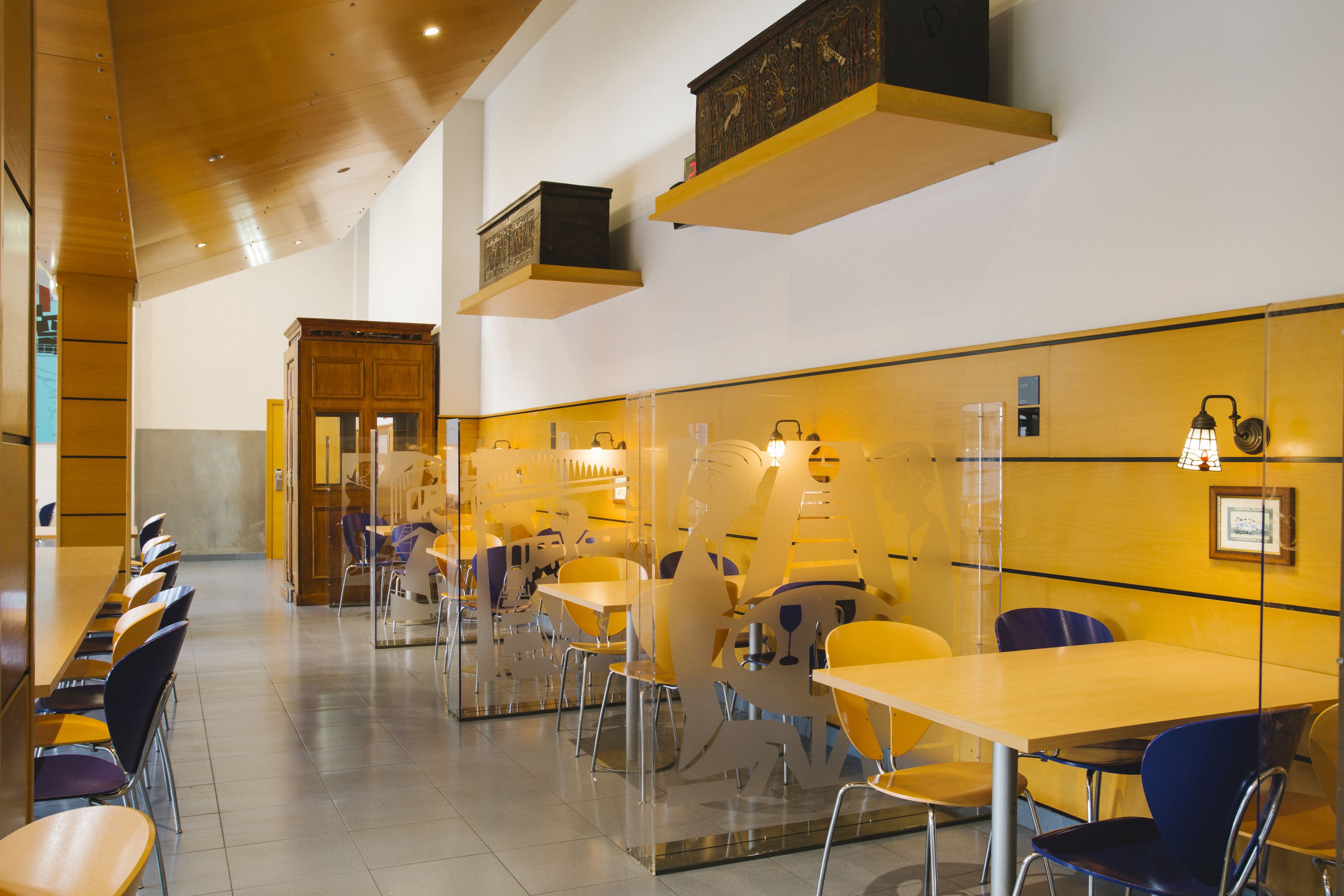 Een moderne eetruimte met lange tafels en stoelen, heldere glazen scheidingswanden en decoratieve wandlampen.