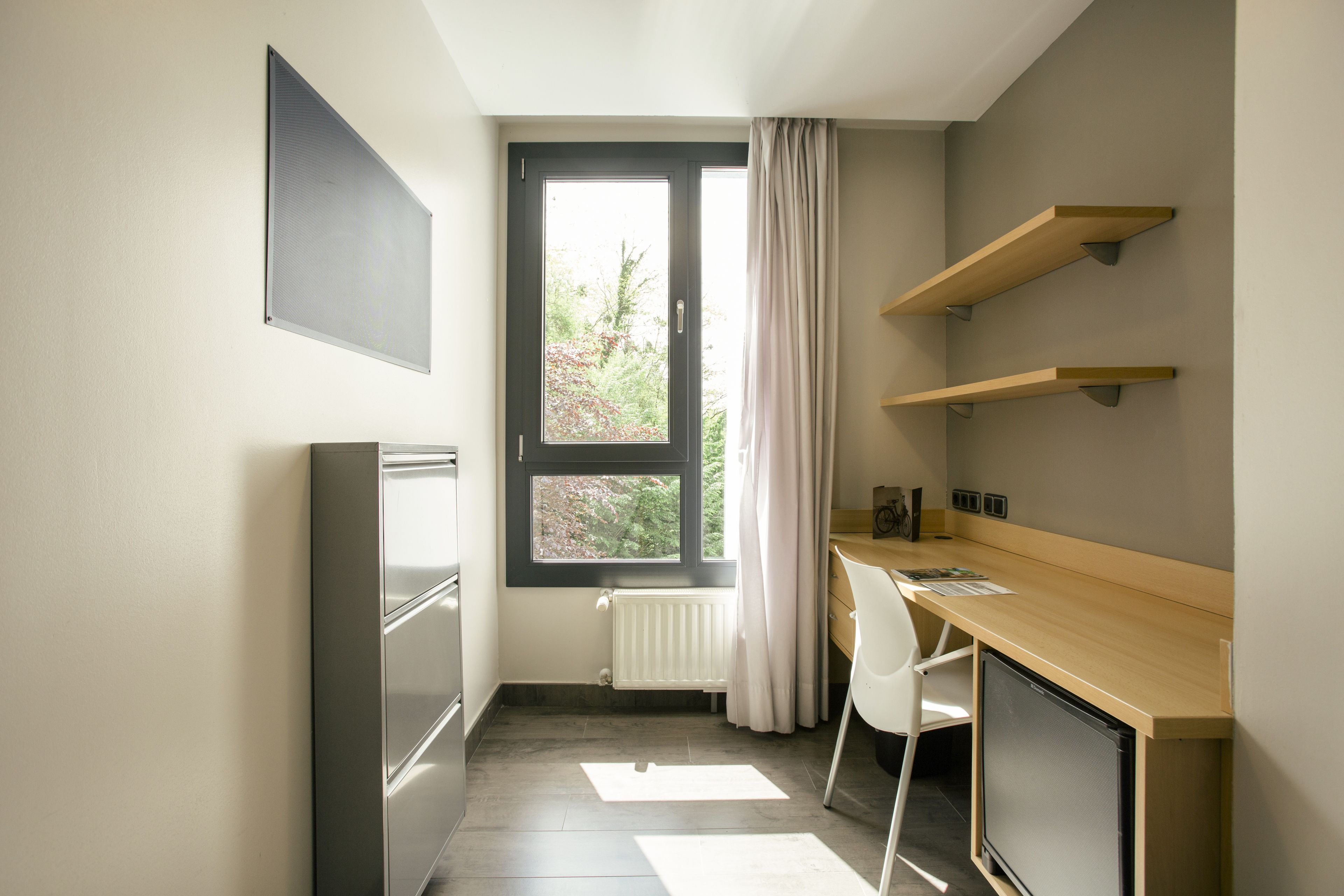 A small, modern kitchen with a refrigerator, a wooden desk, and a window with curtains.