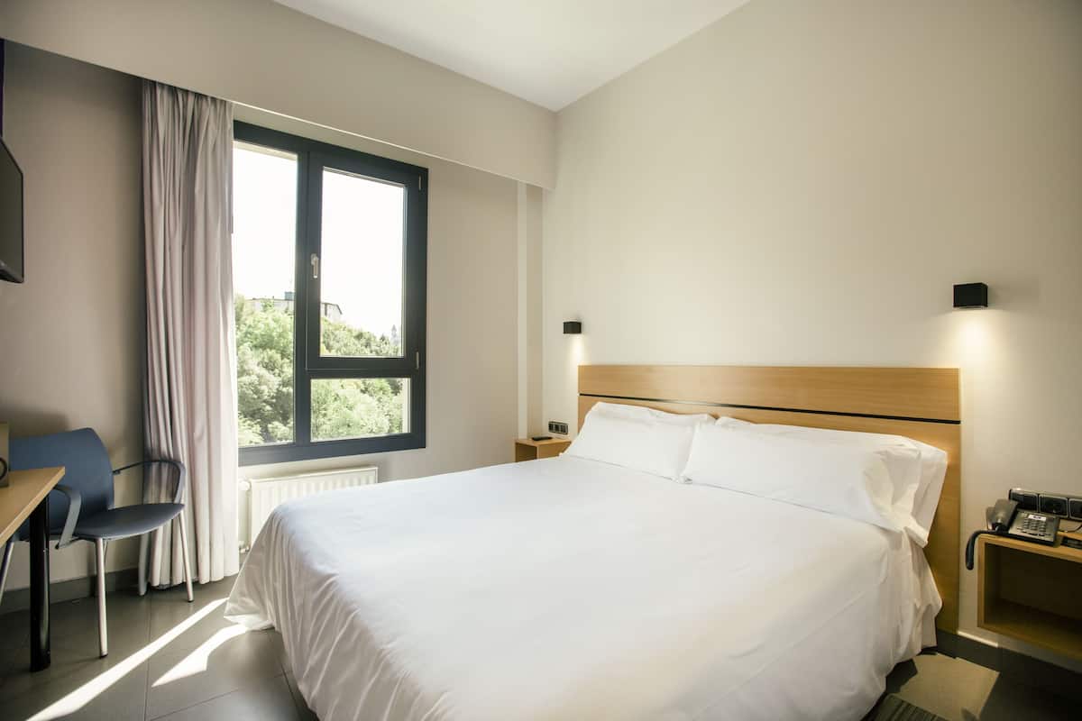 A hotel room with a large bed, a desk with a chair, a window with curtains, a bedside table with a lamp, and a wall-mounted telephone.