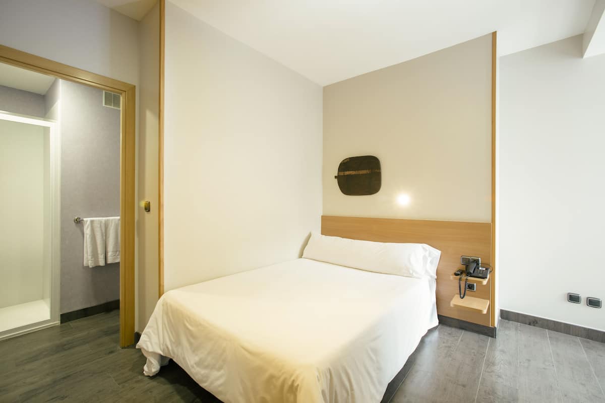 A hotel room with a bed, a wooden headboard, a bedside table with a phone and a lamp, and a towel rack with two towels.