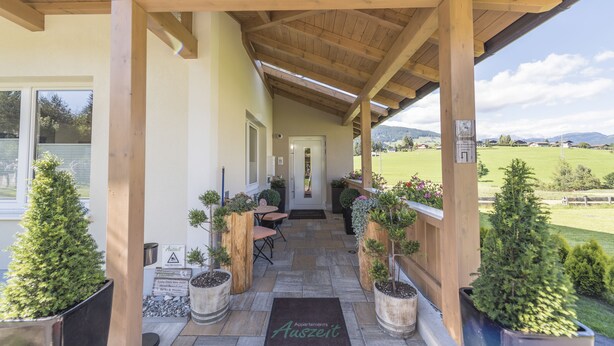 A covered outdoor area with wooden beams, potted plants, a door, and a view of a green landscape. - Appartements Auszeit (Lend)