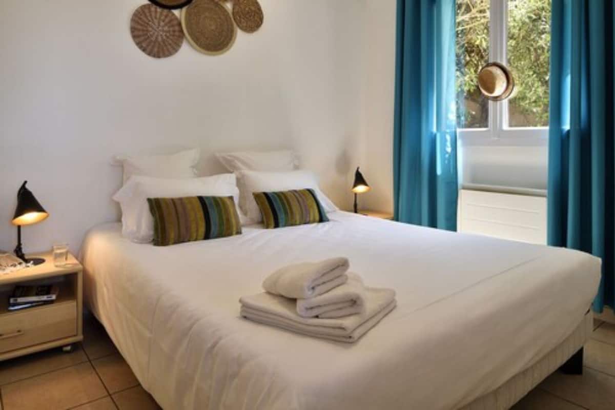 Cama bem arrumada com lençóis brancos e uma pilha de toalhas dobradas, mesa de cabeceira com abajur e janela com cortinas azuis.