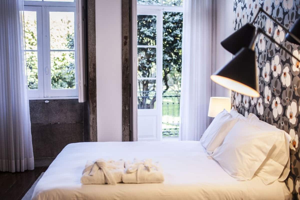 A neatly made bed with white linens and a set of towels, a black lamp, and a window with curtains.