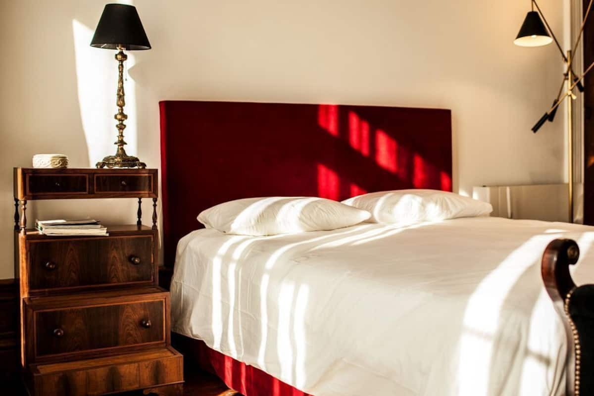 A neatly made bed with white linens, a wooden nightstand with drawers, a red headboard, and two wall-mounted lamps.