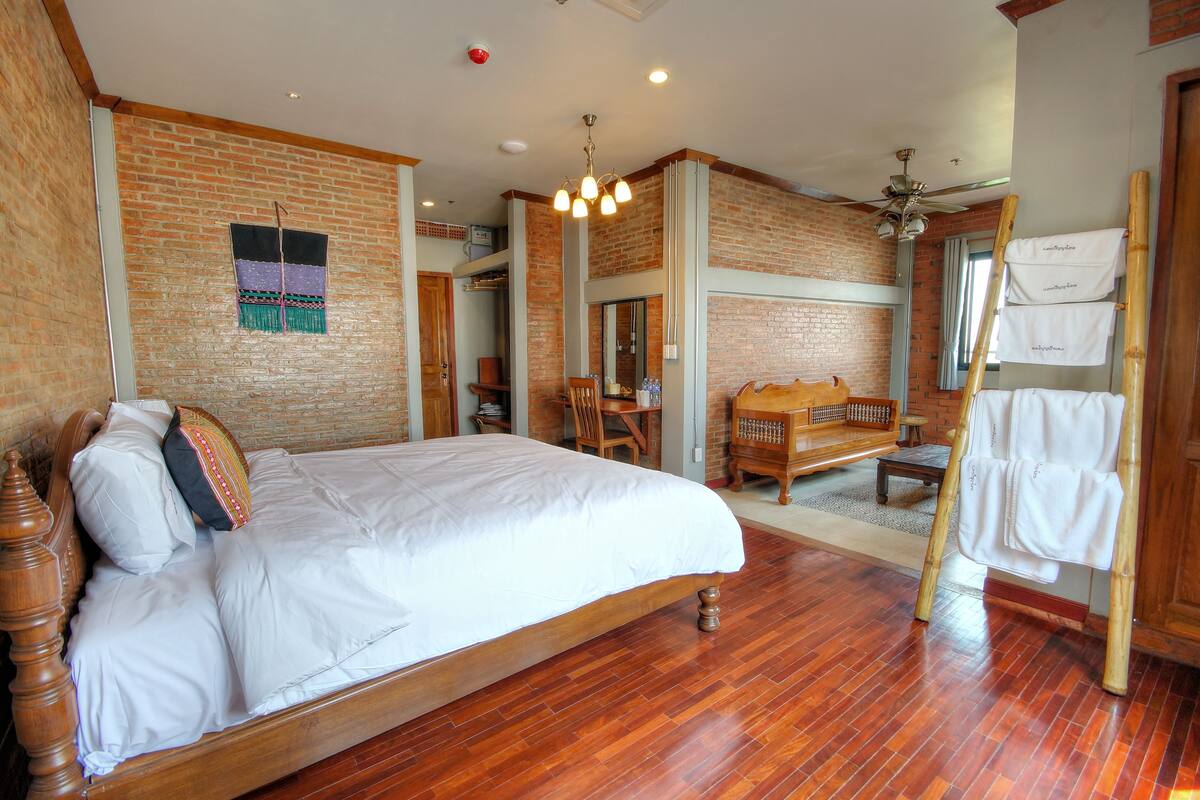 A bedroom with a wooden bed, brick walls, a wooden floor, a ceiling fan, and a wooden dresser with a towel hanging on it.