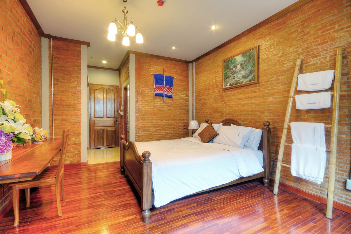 A bedroom with a wooden bed, a dining table with chairs, a brick wall, and a painting.