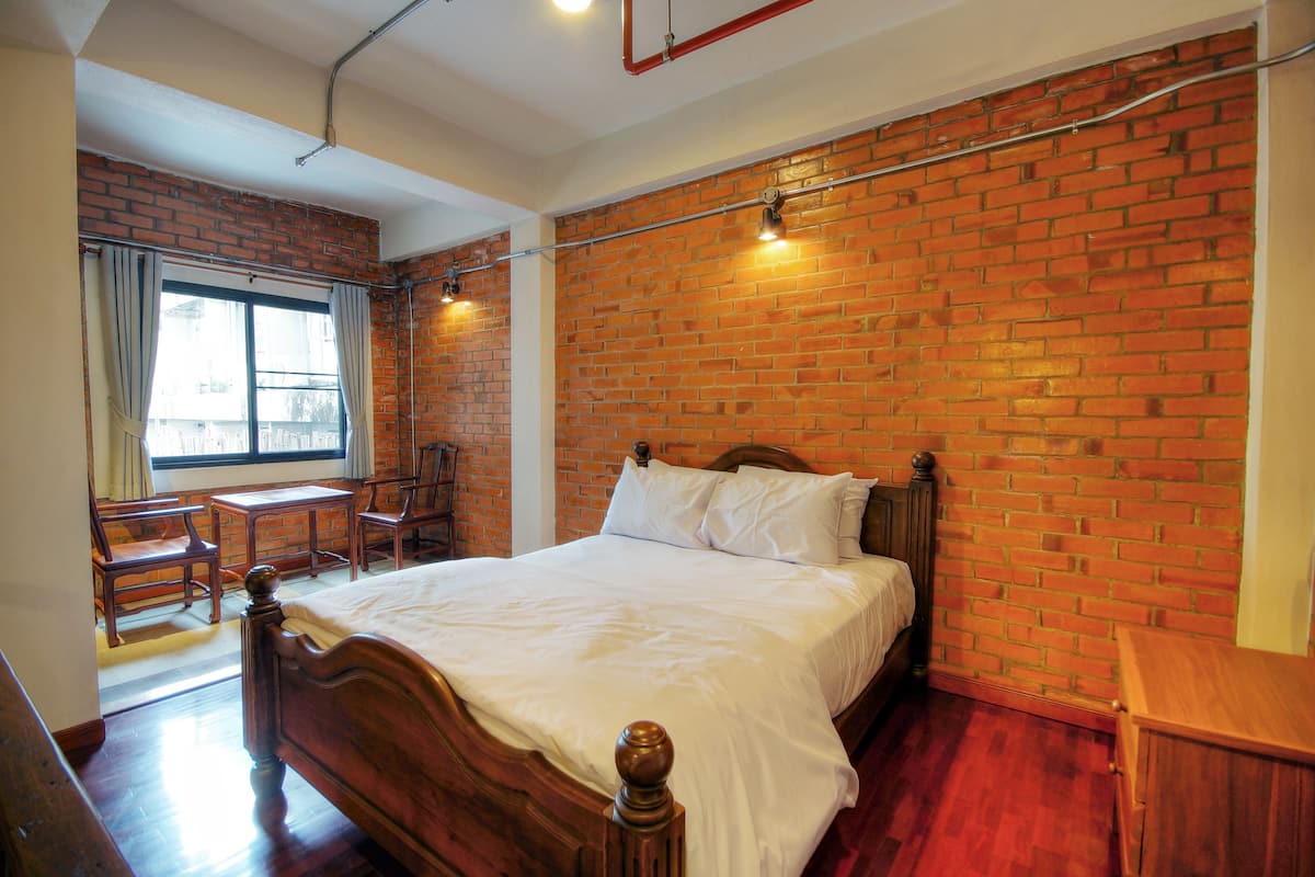 A bedroom with a brick wall, a wooden bed, a window with curtains, and a desk with a chair.