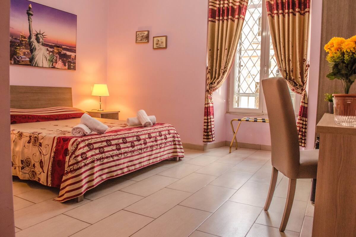A hotel room with a bed, a desk, a chair, a lamp, curtains, and a window with a view of the cityscape.