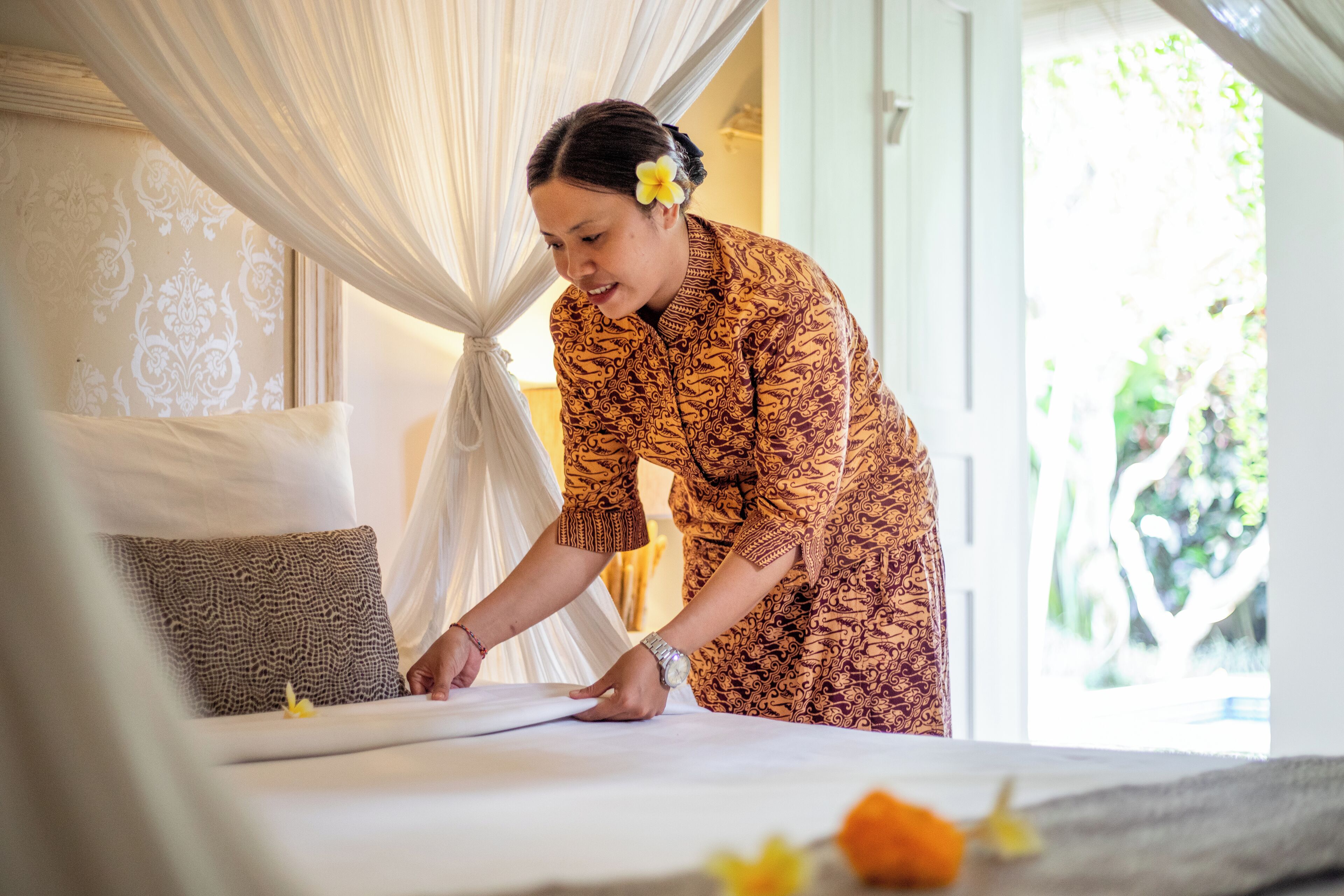 A person arranging a bed with a white and brown patterned bedspread, a white canopy, and a view of greenery outside.