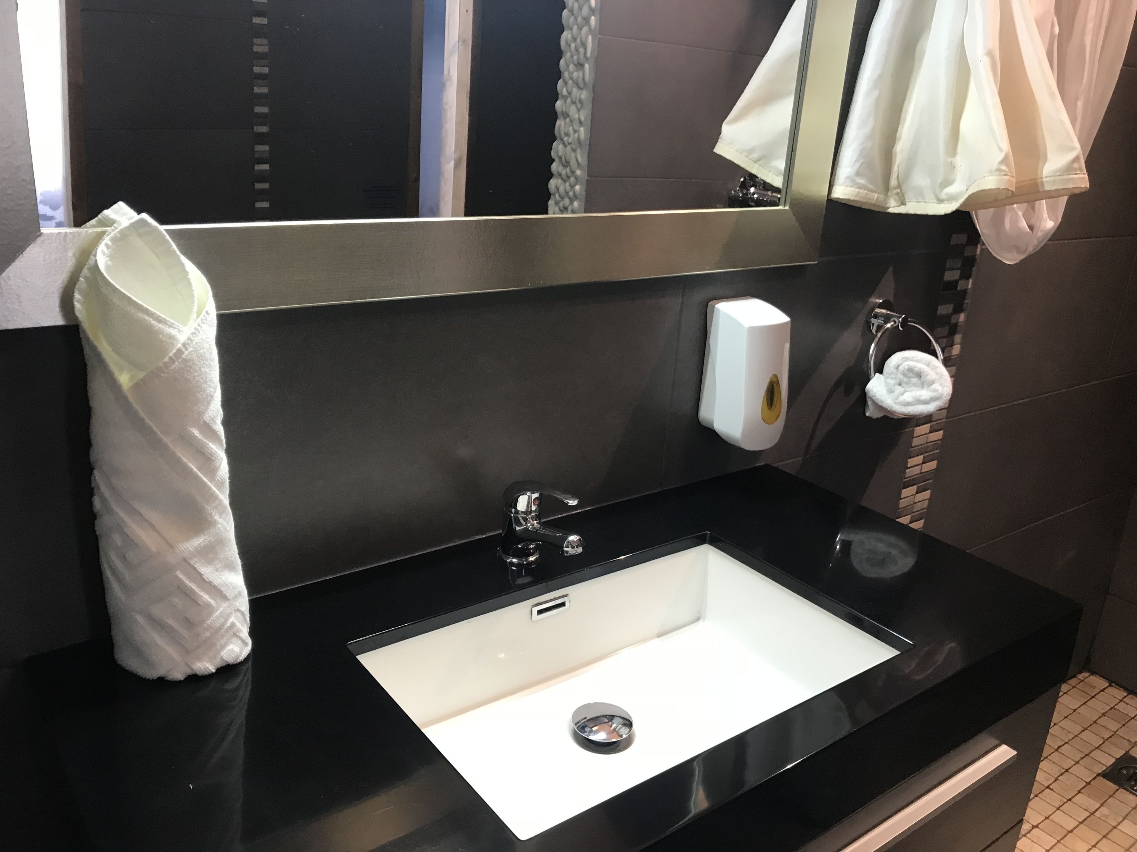 A modern bathroom with a black countertop, a white sink, a mirror, a towel dispenser, and a towel hanging on a rack.