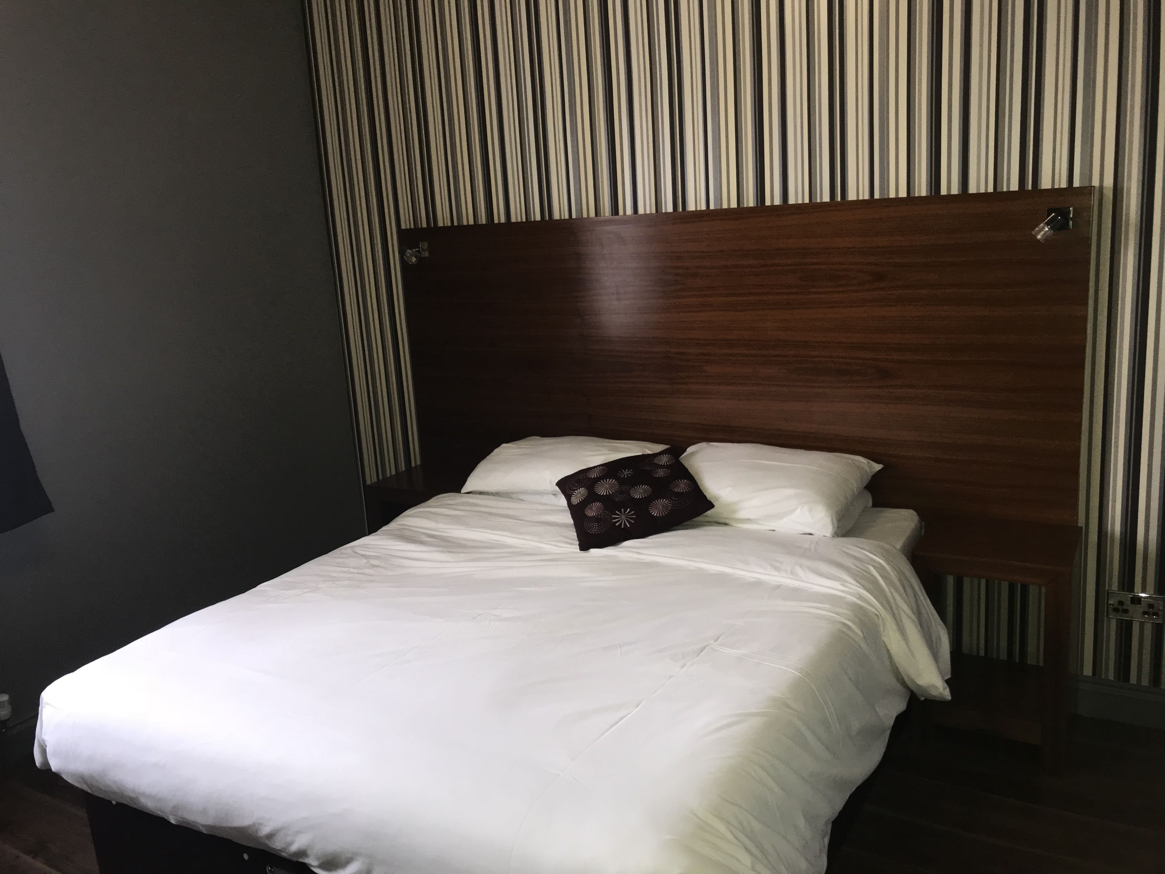 Een net opgemaakt bed met witte lakens en een decoratief kussen, voor een behang met strepen.