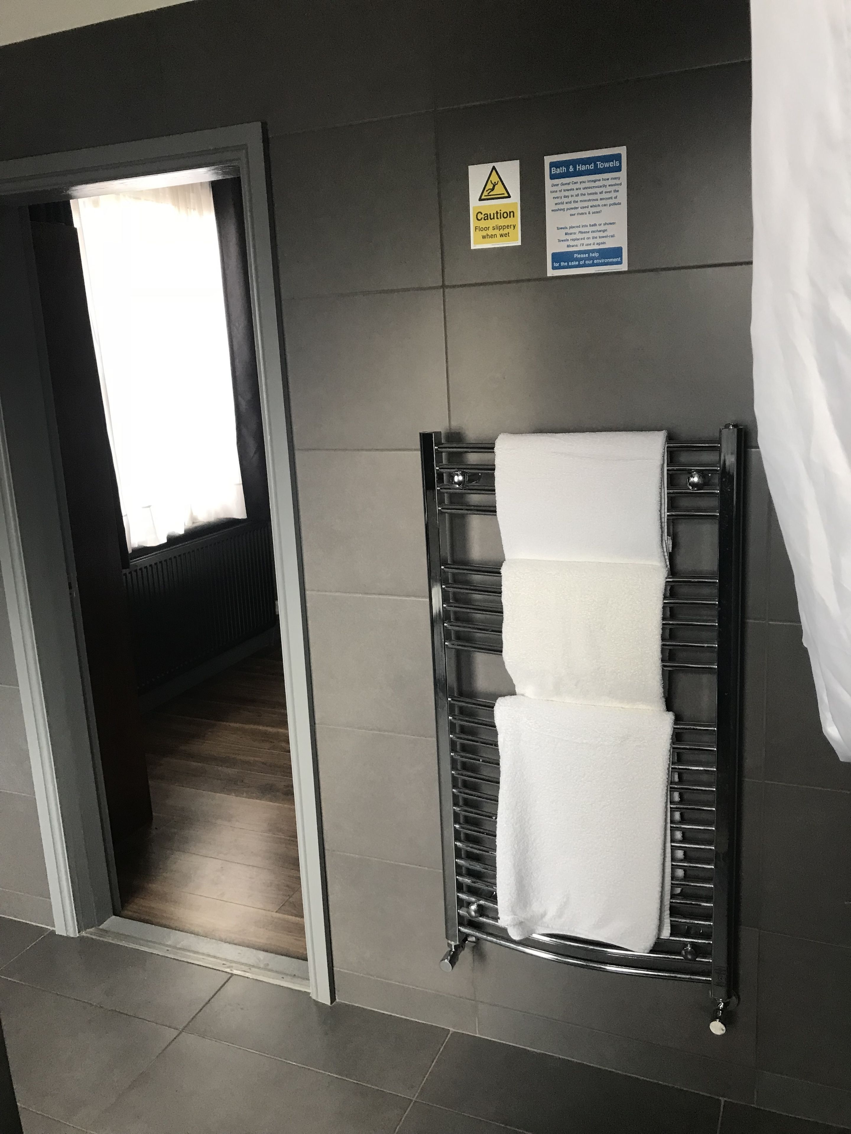 A bathroom with a towel rack holding three white towels, a caution sign for a slippery floor, and a sign offering bath and hand towels.