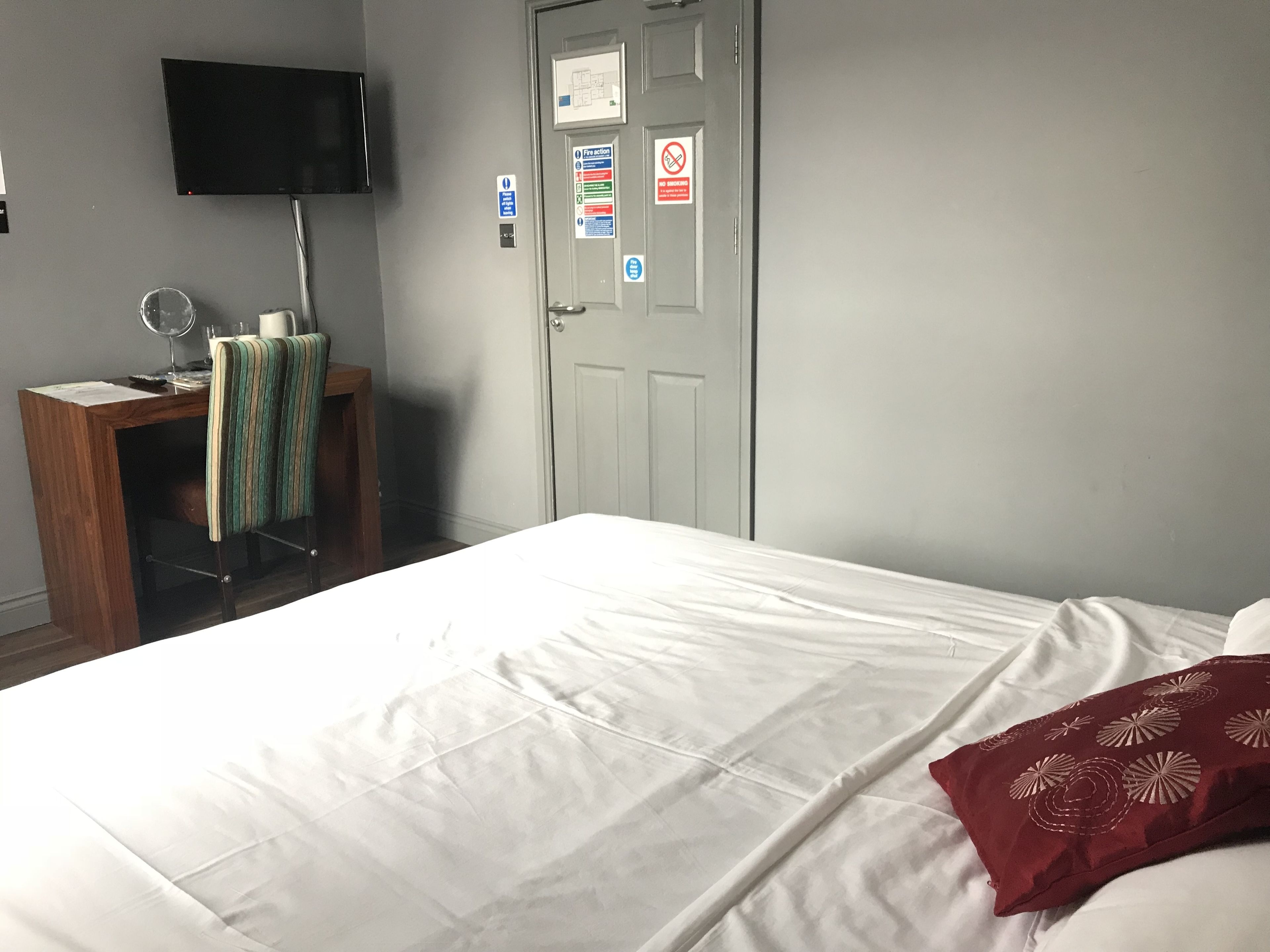 A hotel room with a bed, a desk with a chair, a television, and a door with various signs.