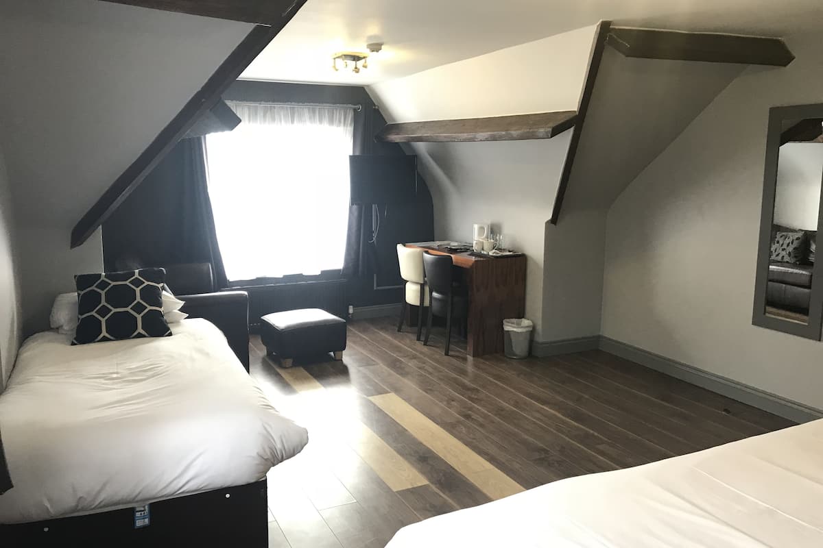 A small, cozy room with a bed, a desk, and a chair. There is a window with curtains, a radiator, and a mirror on the wall.