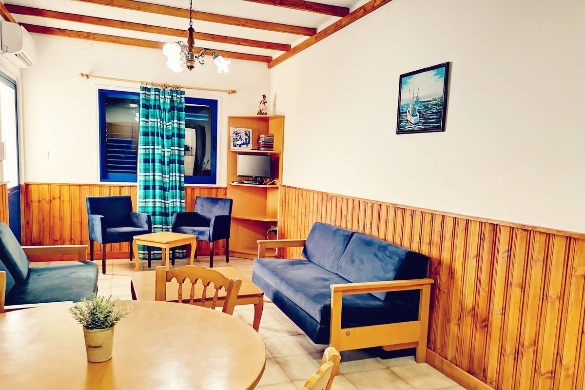 A cozy living room with a wooden ceiling, a blue sofa, a wooden dining table, and a television on a wooden shelf.