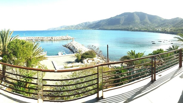 A coastal view from a balcony with a railing, a rocky area with a small pool, and a mountain in the background. - Kanalli Restaurant & Apartments (Pomos)