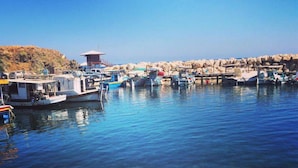 A marina with boats docked, clear water, and rocky terrain in the background. - Kanalli Restaurant & Apartments (Pomos)