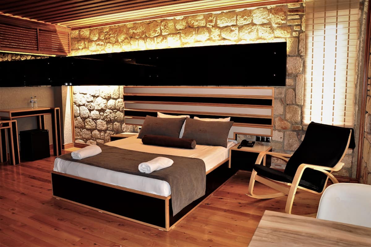 A modern bedroom with a stone wall, a large bed with pillows, a wooden nightstand, a black chair, and a wooden desk.