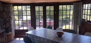 Un coin repas avec une table dressée pour six personnes, de grandes fenêtres donnant sur un paysage verdoyant et une plante en pot posée sur un tronc d’arbre.