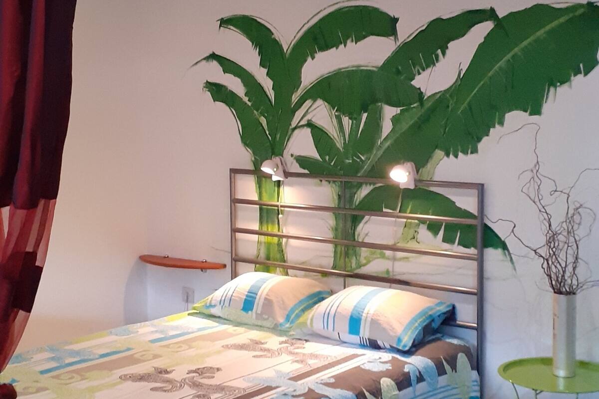 Een slaapkamer met een bed, een grote kunstplant, een klein tafeltje en een gordijn.