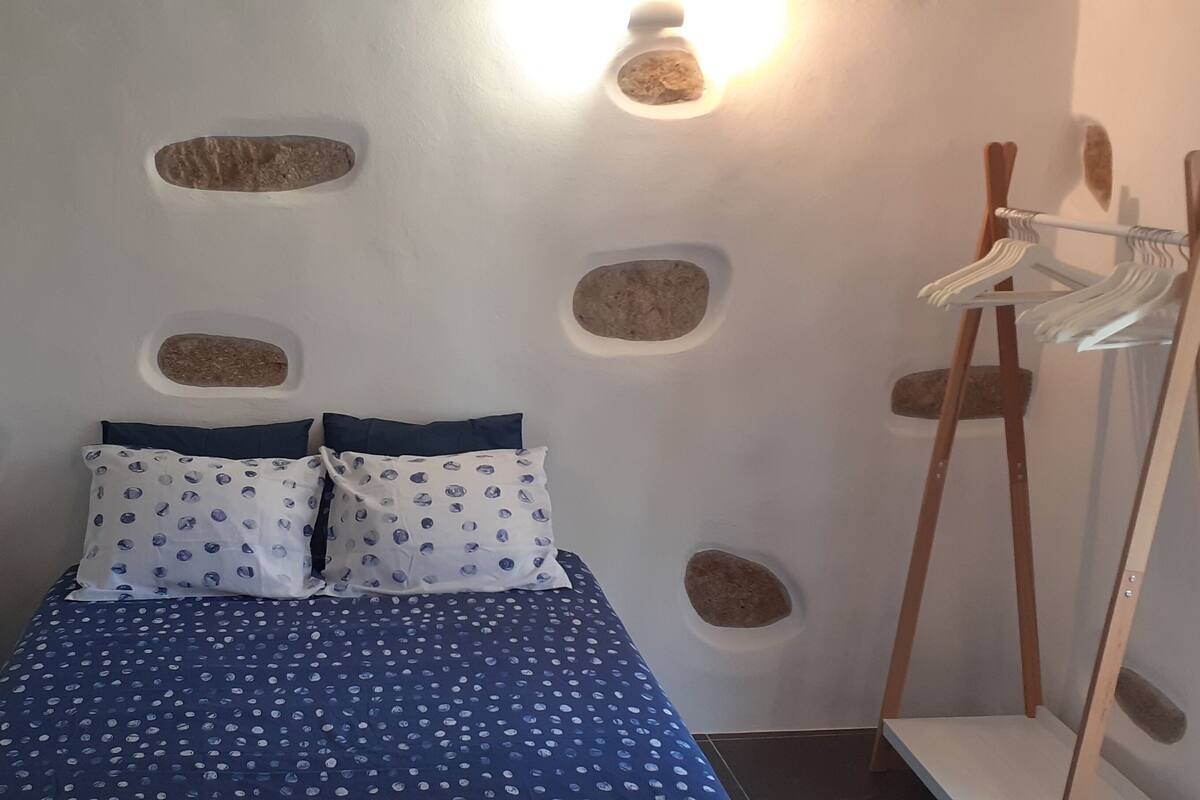 Een slaapkamer met een bed, twee kussens, een stoel, een houten kapstok en rotsen aan de muur.