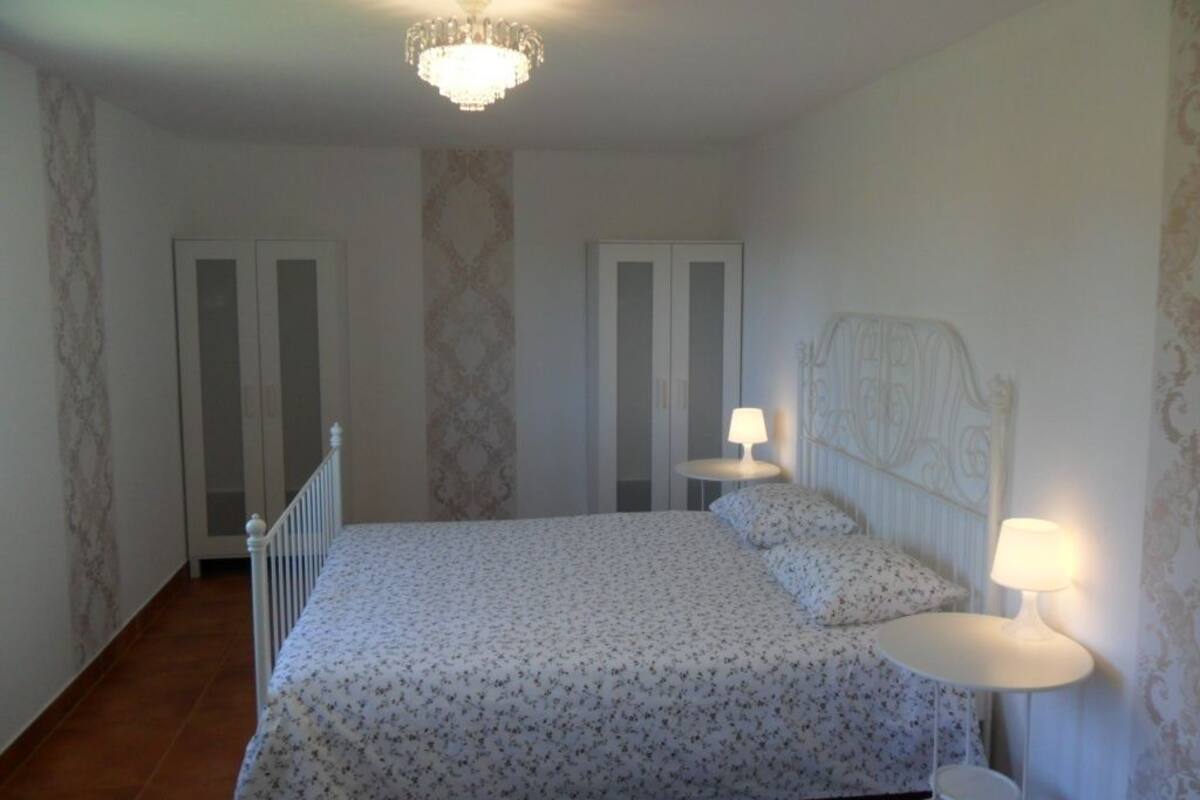 Een slaapkamer met een bed, twee nachtkastjes met lampen, een kroonluchter en een deur.