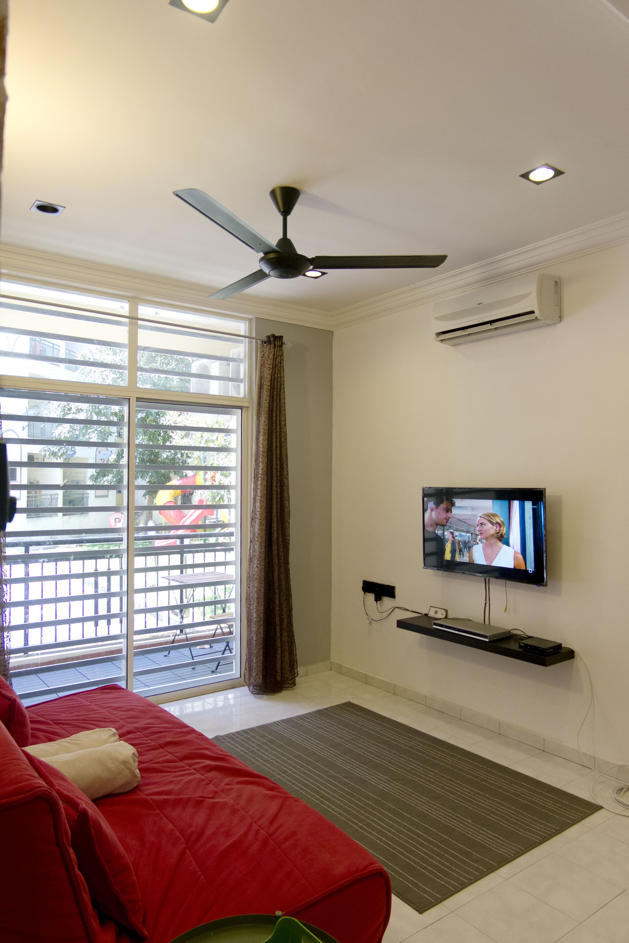 Ein Wohnzimmer mit einem roten Sofa, einem an der Wand montierten Fernseher, einem Deckenventilator und einem großen Fenster mit Jalousien.