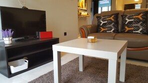Una sala de estar moderna con un televisor de pantalla plana, una mesa de centro blanca, un sofá con cojines estampados y una planta decorativa.