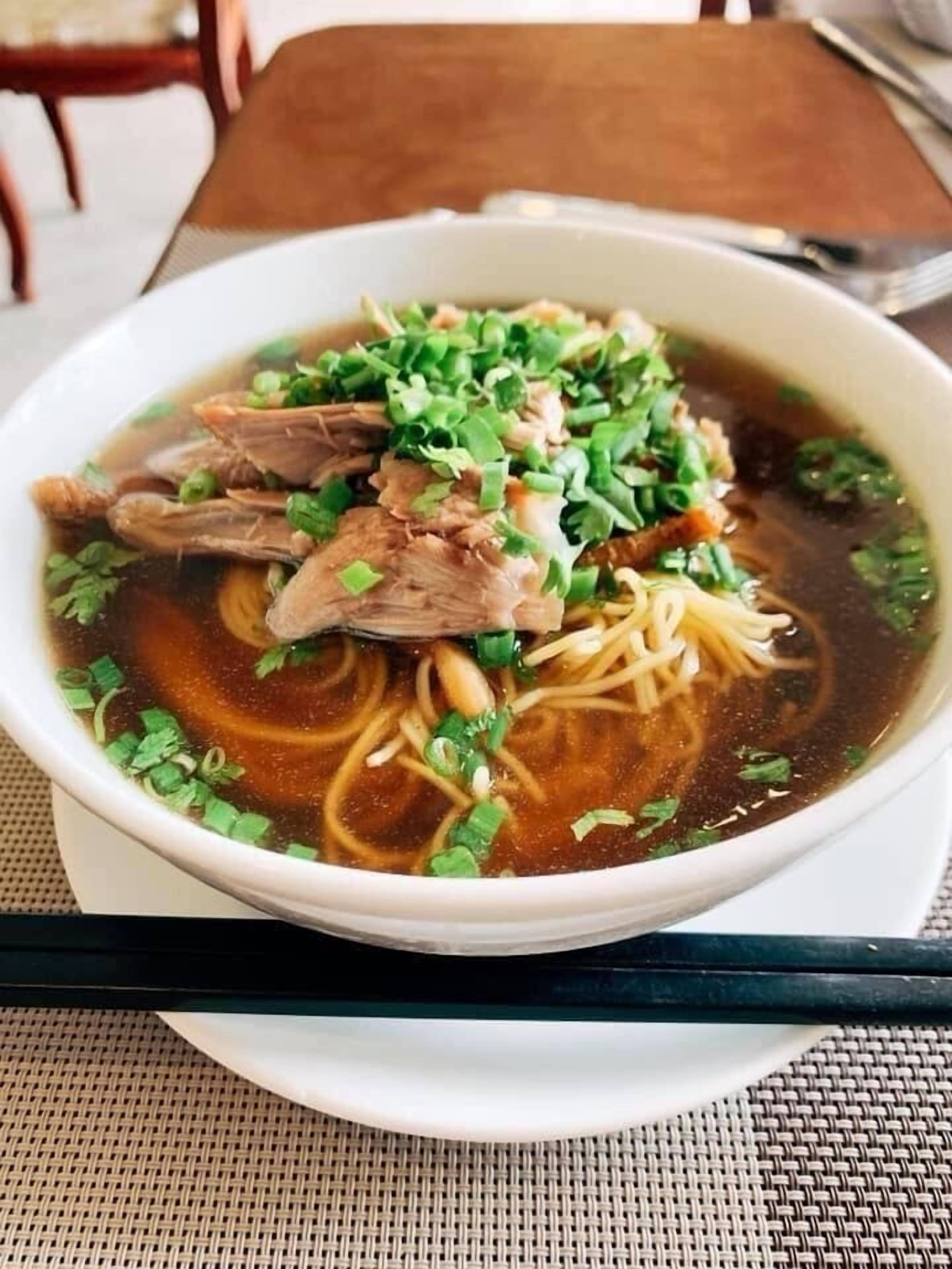 Eine Schüssel Nudelsuppe mit Fleisch und Frühlingszwiebeln, serviert in einer weißen Schüssel auf einem Tisch mit Untersetzer aus Bändern.