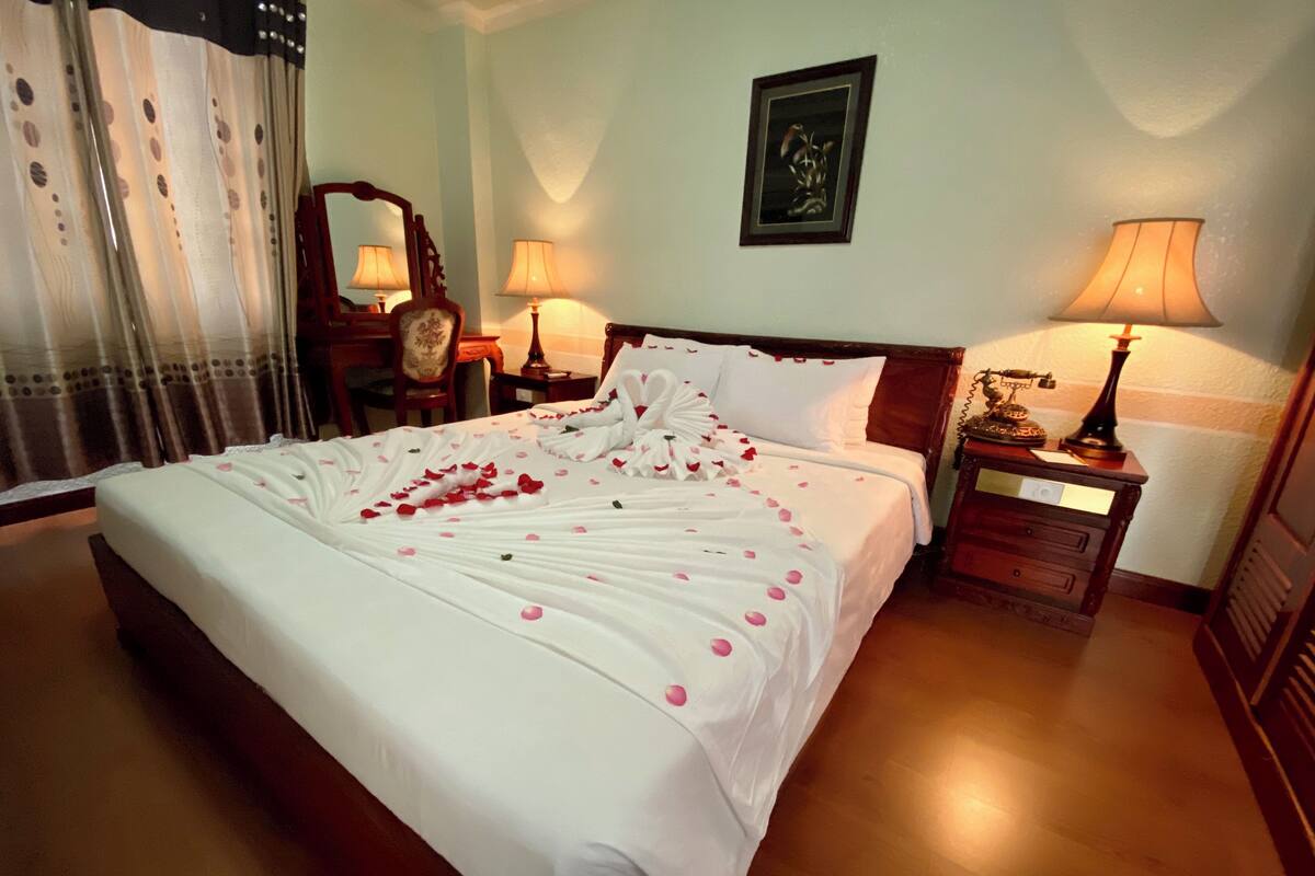 Una cama bien hecha con pétalos de rosa, dos lámparas de noche, un cuadro en la pared y una mesita de noche de madera con un teléfono.