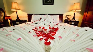 Una cama con sábanas blancas y pétalos de rosa roja dispuestos en forma de corazón.