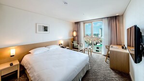 A hotel room with a large bed, a desk, a chair, a TV, and a window with a city view. - Jeju Central City Hotel (Jeju-si,)
