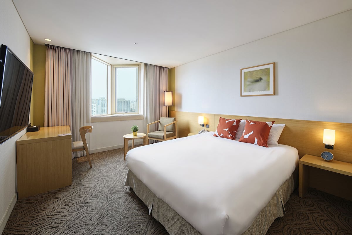Une chambre d’hôtel avec un grand lit, un bureau, une chaise, une petite table, une table de chevet, une horloge et une fenêtre avec des rideaux.