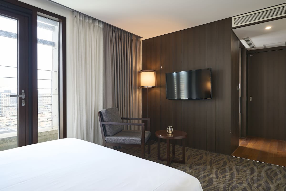 Une chambre d’hôtel avec un lit, une chaise, une petite table, un téléviseur fixé au mur et une vue sur la ville par la fenêtre.