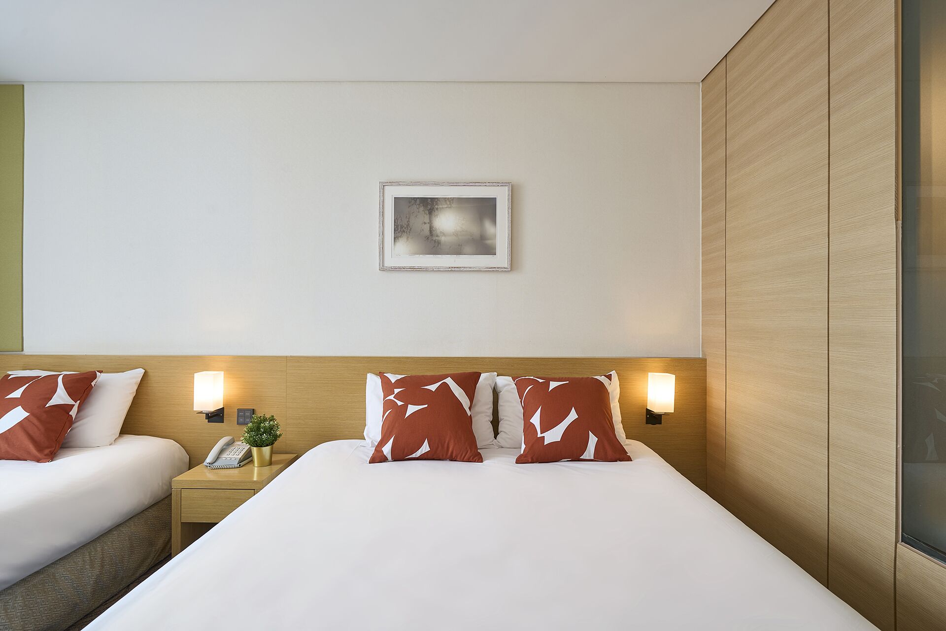 Quarto de hotel com duas camas, uma mesa de cabeceira com abajur, uma pequena planta e um quadro na parede.
